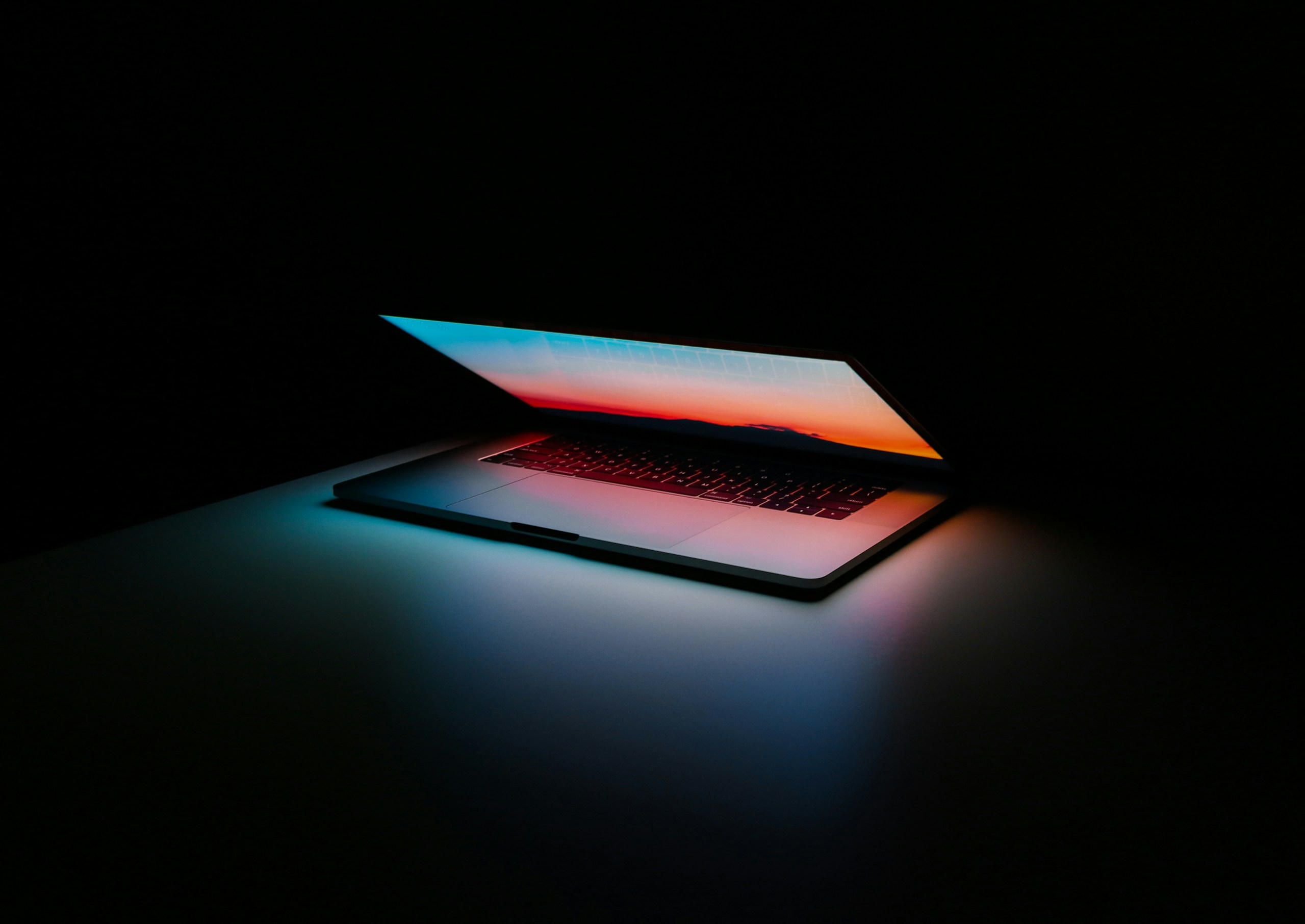 A laptop opening in a dark setting, light from the screen casting dramatic color across the scene